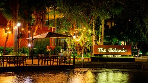 An outdoor seating area with tables and chairs, a sign for 'The Botanic service room', and a water feature in the foreground. - The Botanic Service Room (Pak Kret)