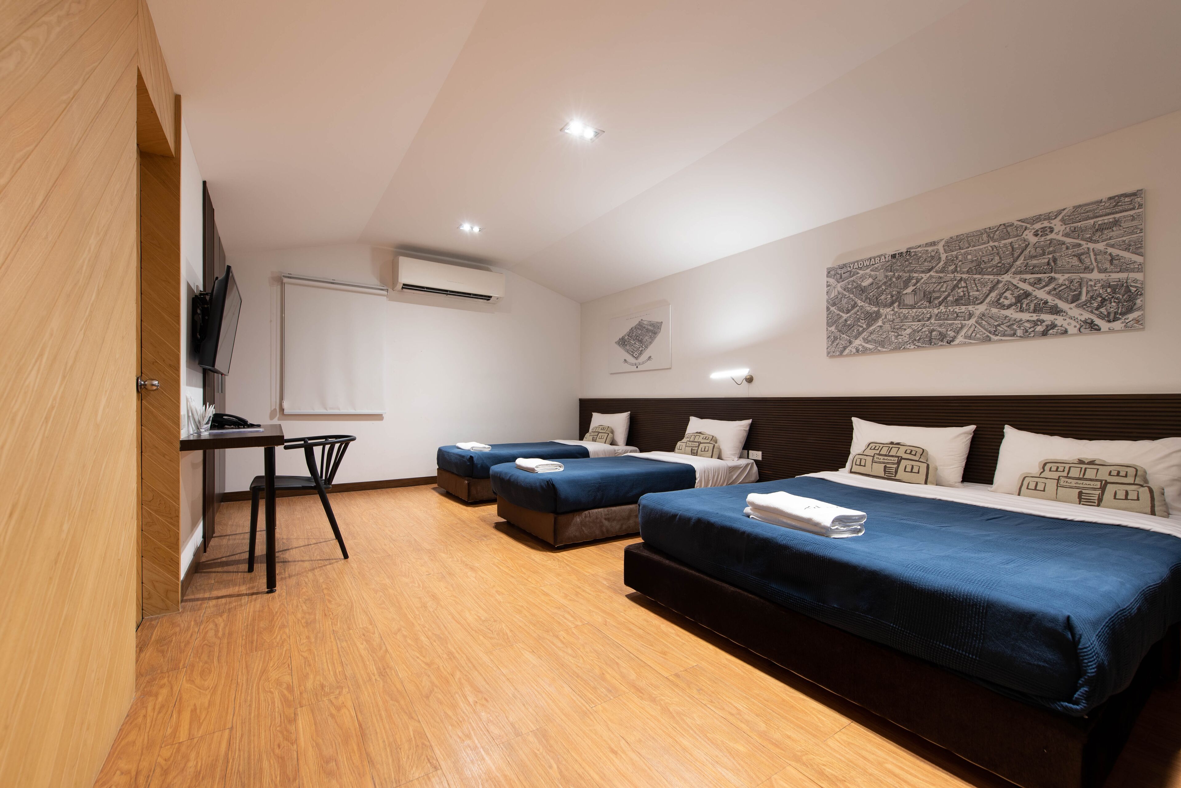 A hotel room with a wooden floor, a desk with a chair, two single beds, a double bed, and a wall-mounted TV.