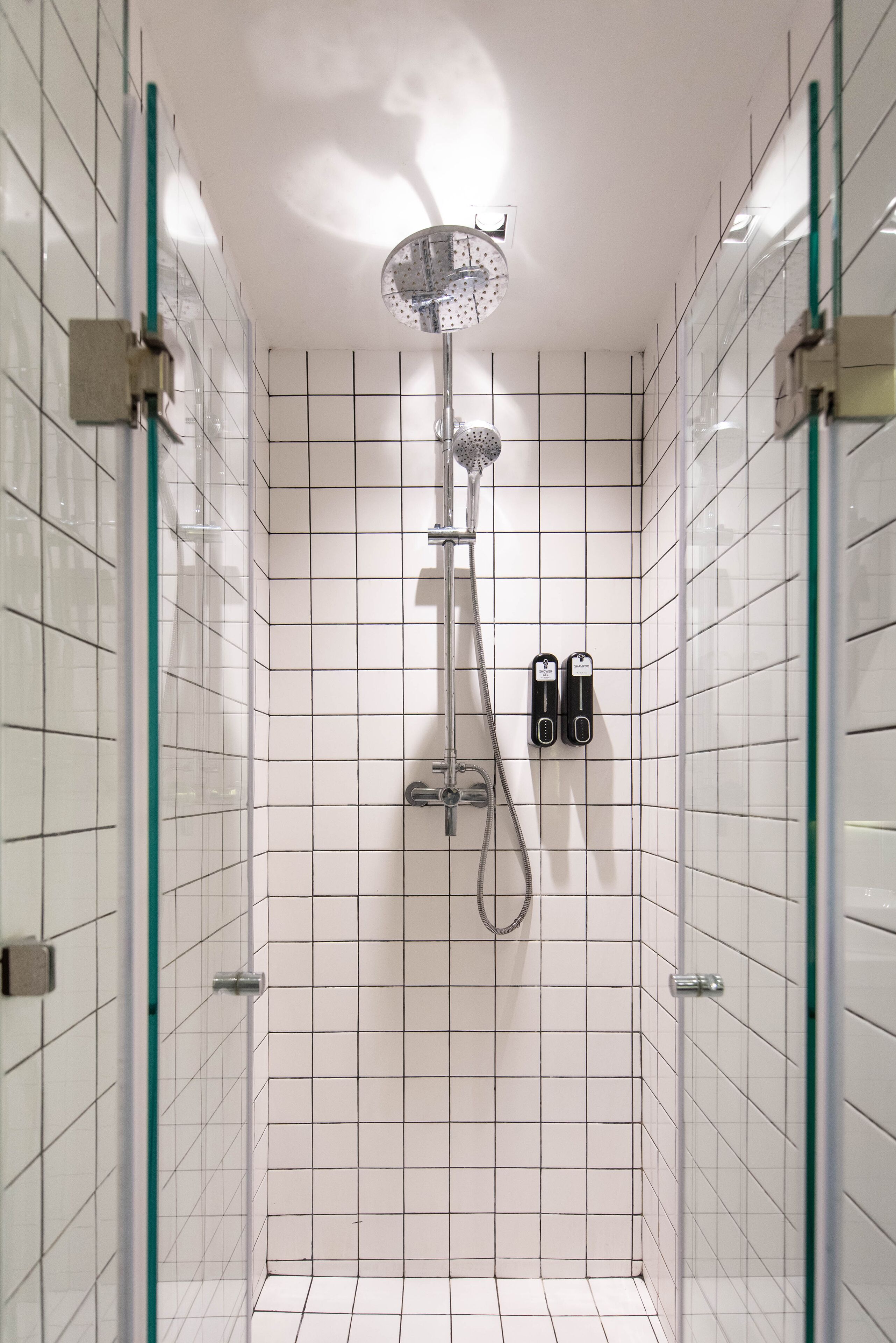 Et moderne brusebad med glasdør, hvide flisevægge og et håndholdt brusehoved.