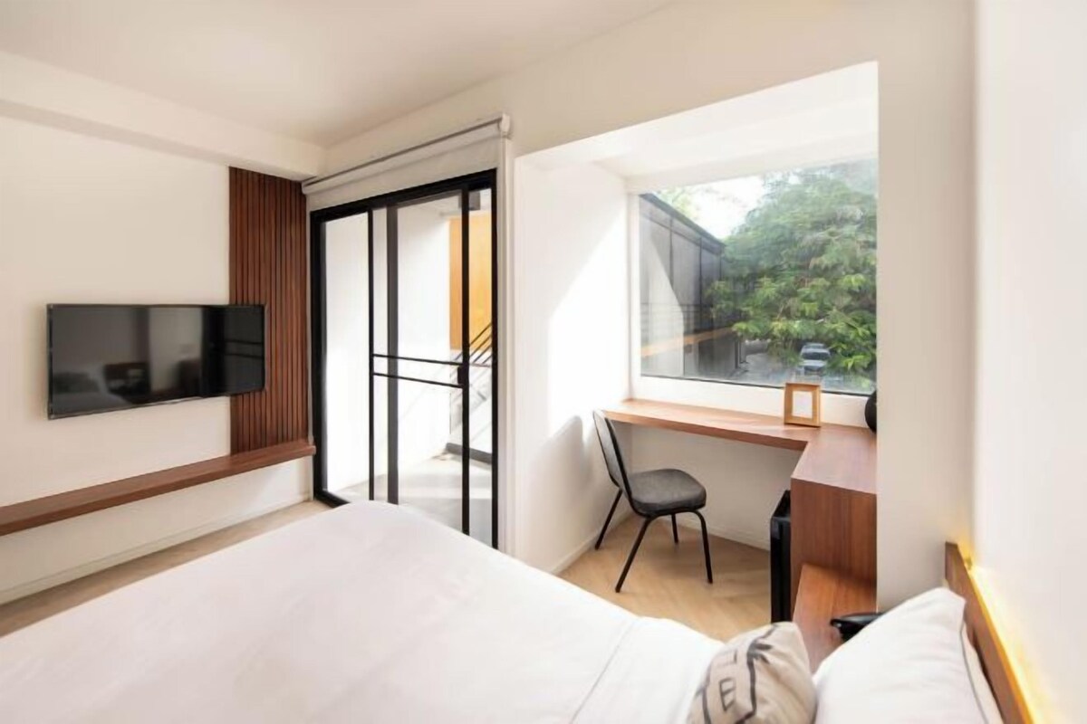 A modern hotel room with a large bed, a desk with a chair, a TV mounted on the wall, and a window offering a view of greenery outside.