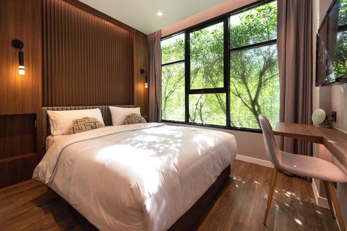 Ein modernes Schlafzimmer mit einem großen Bett, einem hölzernen Kopfteil, einem Schreibtisch mit Stuhl und einem großen Fenster mit Blick auf Bäume.