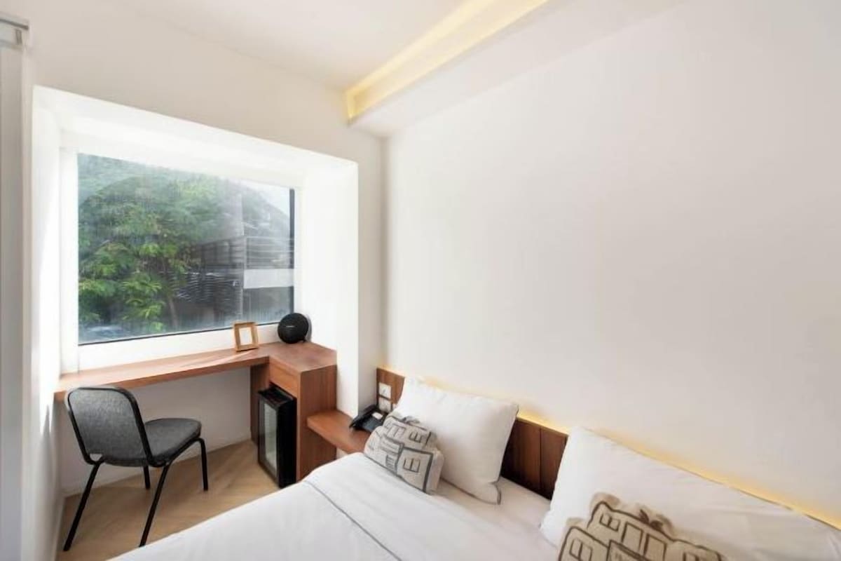 A bedroom with a bed, a desk, a chair, and a window with a view of greenery.