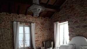 Un dormitorio con paredes de piedra, techo de madera, lámpara de araña, cama con ropa blanca y una ventana con cortinas.