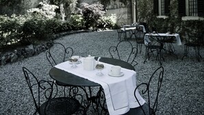 Un espacio de comedor al aire libre con mesas y sillas, rodeado por un sendero de grava y un muro de piedra.
