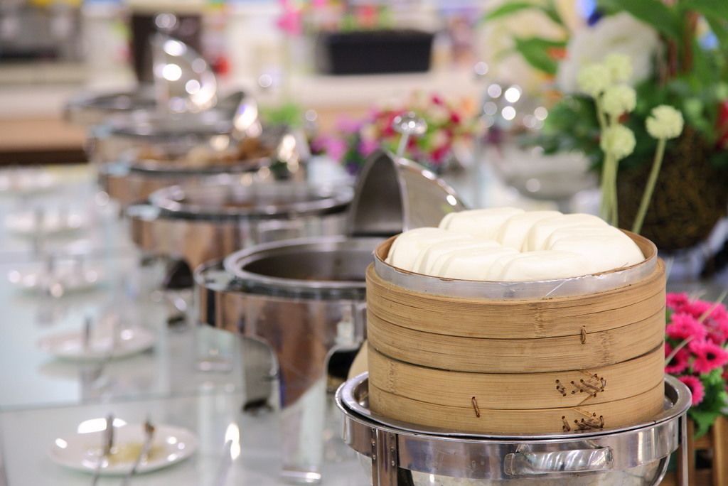 A buffet setup with multiple food containers, including a prominent bamboo steamer with white food.