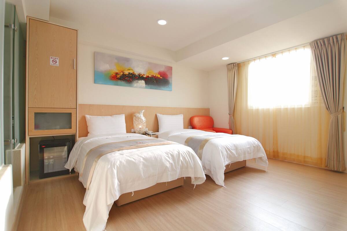 Quarto de hotel com duas camas, um guarda-roupa de madeira, um quadro na parede e uma janela com cortinas.