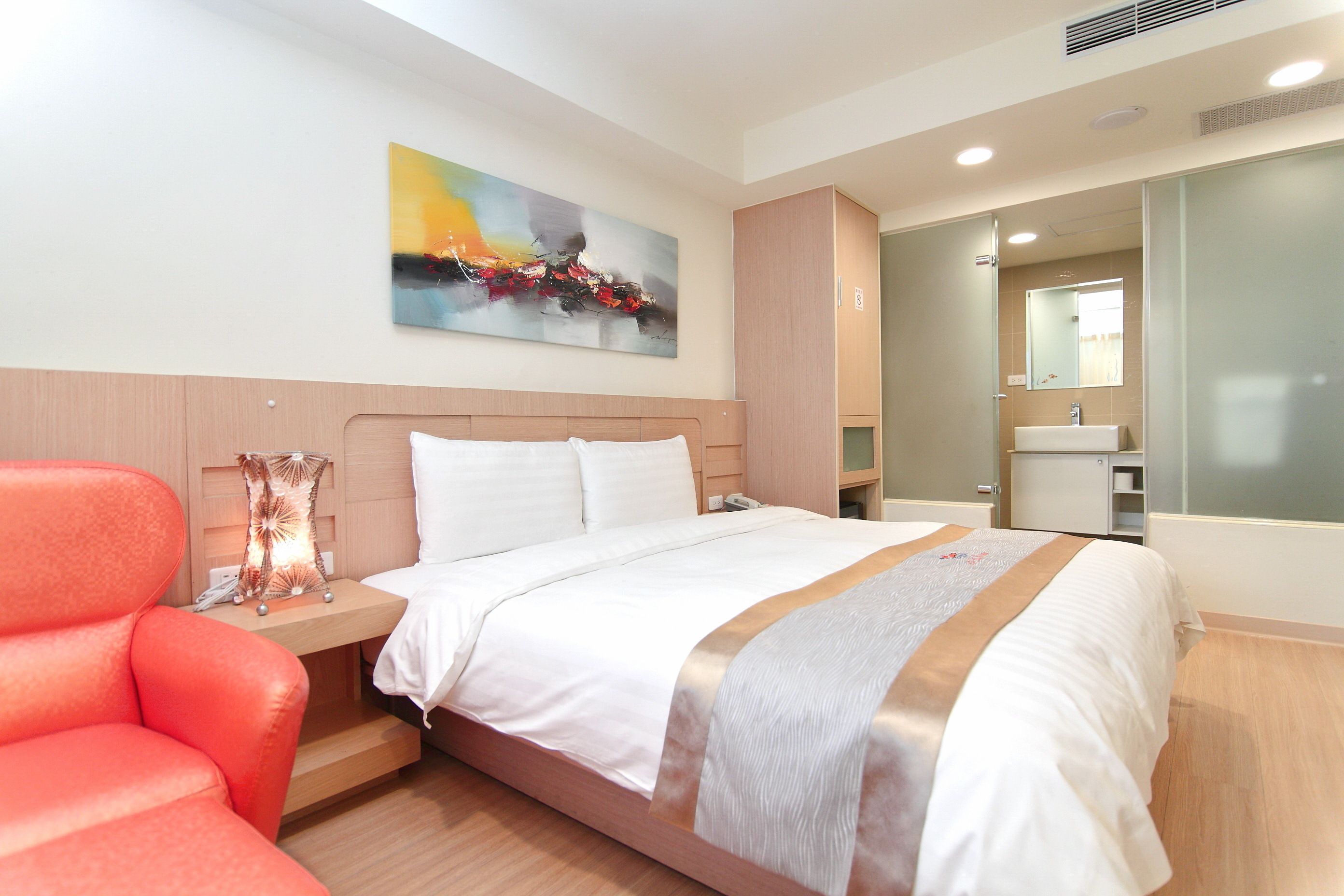A modern hotel room with a large bed, a nightstand, a red chair, a painting on the wall, and a bathroom with a mirror and sink.