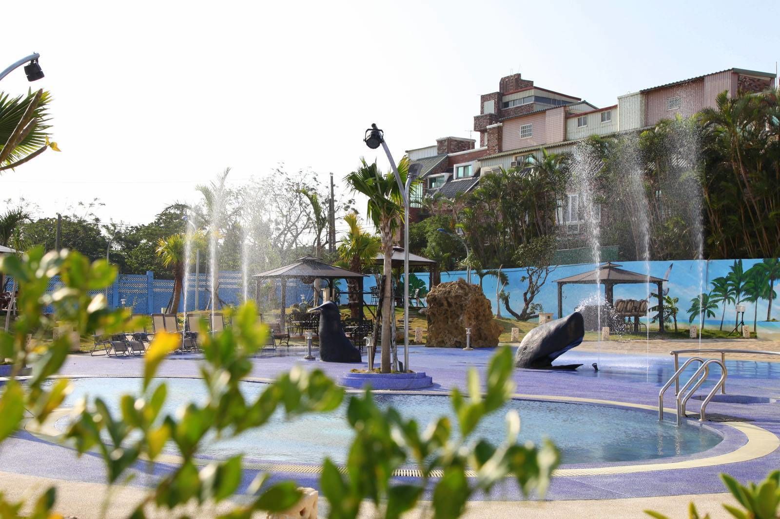 Una fuente con esculturas de ballenas en un estanque, rodeado de palmeras y un edificio al fondo.