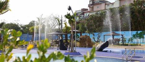 Una fuente con esculturas de ballenas en un estanque, rodeado de palmeras y un edificio al fondo.