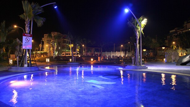 A nighttime swimming pool area with illuminated water, palm trees, and a sign indicating no diving. - Clownfish Resort (Hengchun)