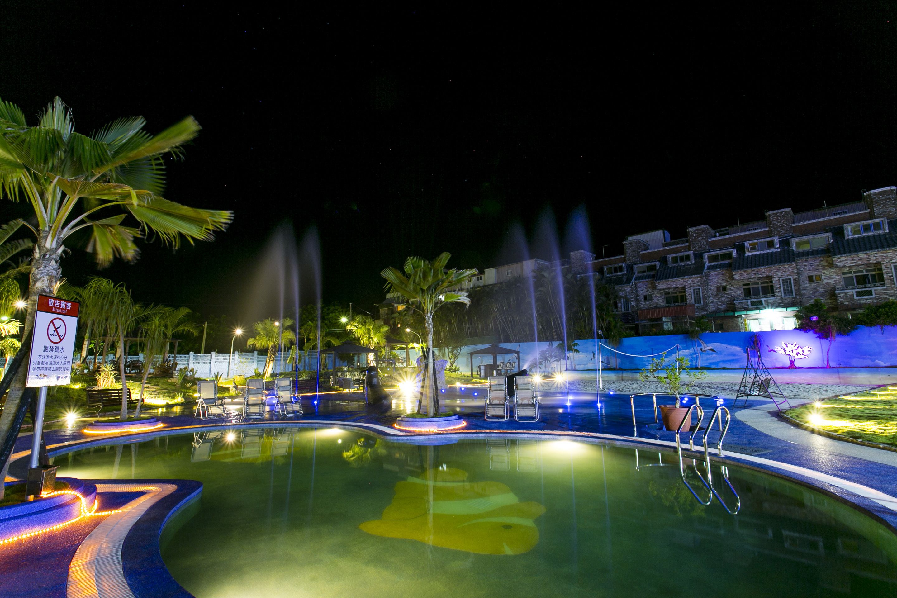 A nighttime swimming pool with water fountains, palm trees, and a building in the background.