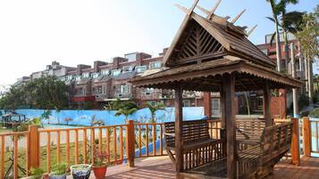 A wooden gazebo with a sloped roof, surrounded by a wooden fence, overlooking a pool area with a blue wall and palm trees.