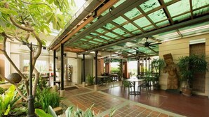 Un espacio al aire libre techado con mesas y sillas, rodeado de plantas y una estatua.