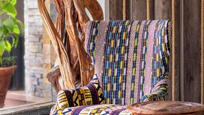 A chair with a colorful geometric pattern, a wooden sculpture, and a small wooden table in a room with a stone wall and a wooden panel. - The Monarch Hotel (Nairobi)