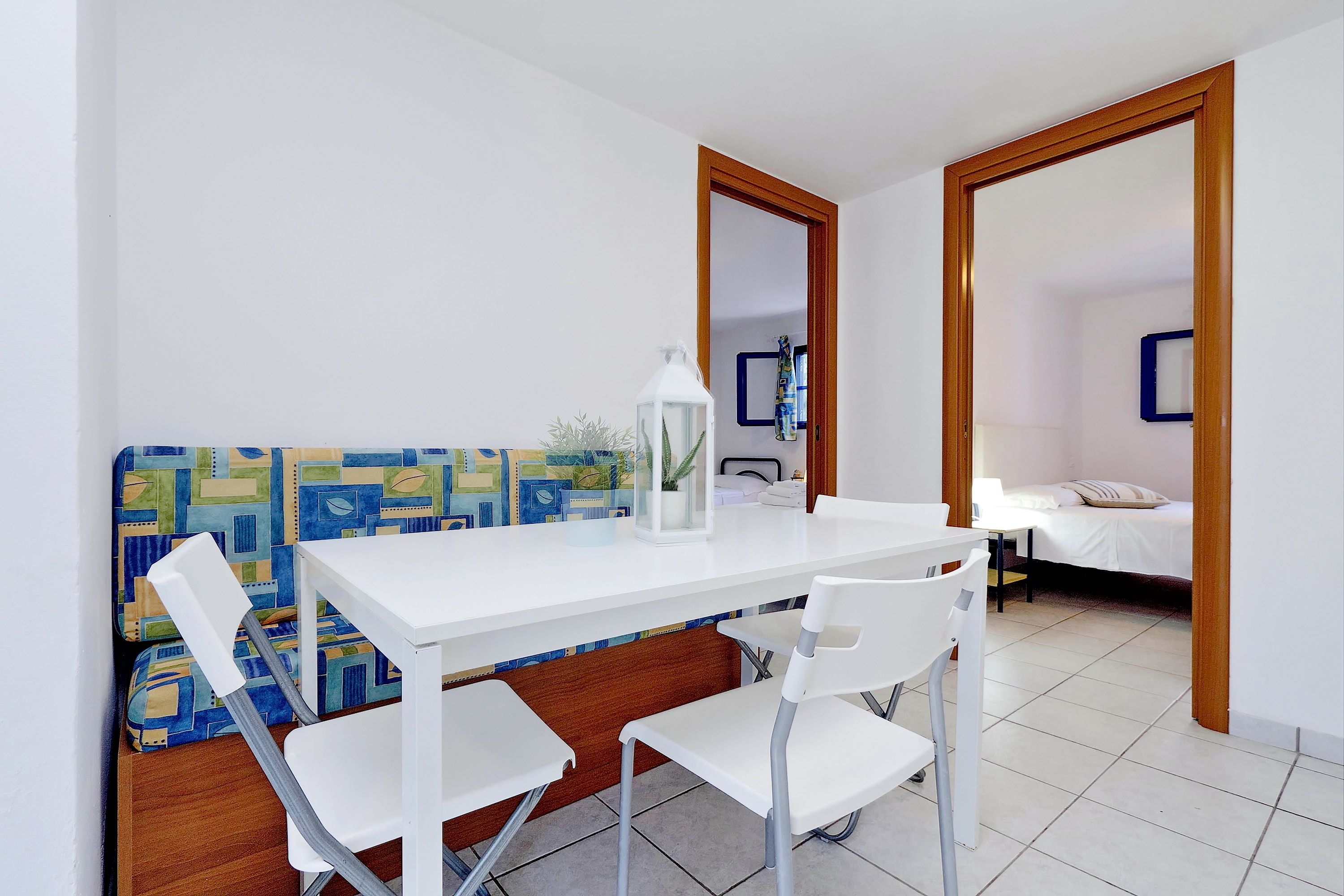 A small, clean room with a white table and chairs, a wooden bench, and a bed with a white mattress and pillow.