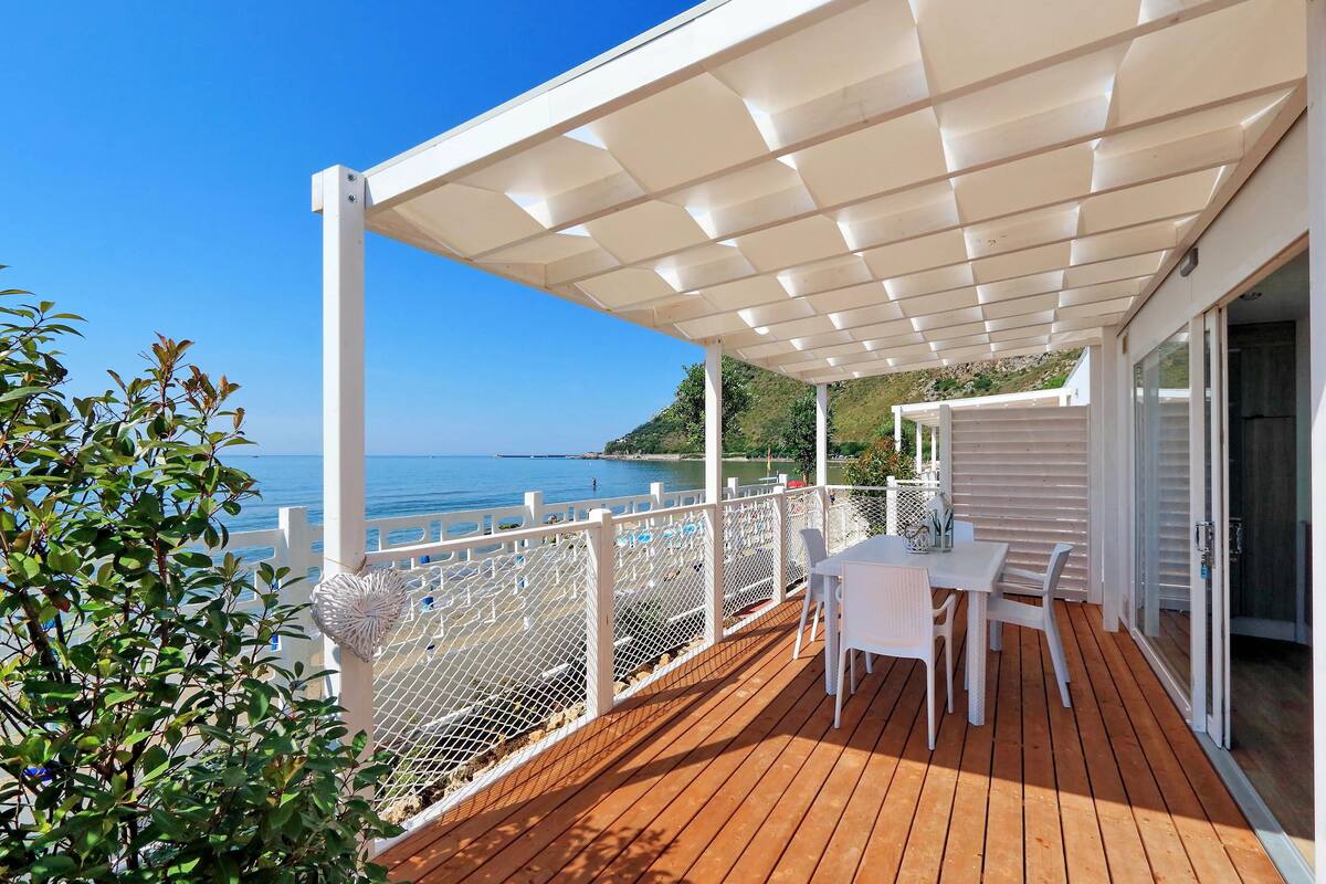 A wooden deck with a white pergola, white chairs, and a table overlooking the sea.