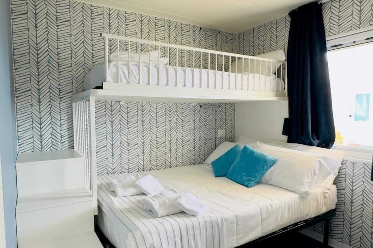 A bunk bed room with a herringbone patterned wallpaper, a white bed with striped bedding, and a window with a black curtain.