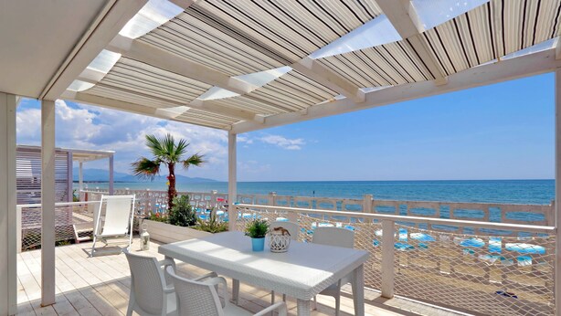 Un espace extérieur abrité, avec une table et des chaises, donnant sur une plage parsemée de parasols et agrémentée d’un palmier.