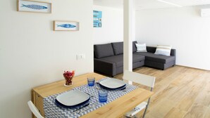Un coin repas avec une table en bois dressée pour deux personnes, un canapé gris et une petite table d’appoint sur laquelle est posé un vase de fleurs.