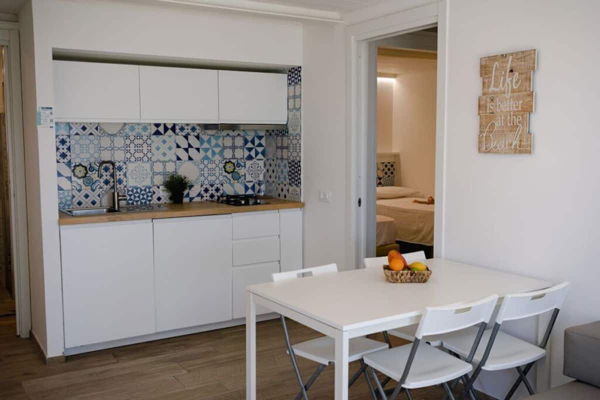 A compact kitchen with white cabinets and blue patterned backsplash, a dining table with chairs, and a small fruit basket on the table.