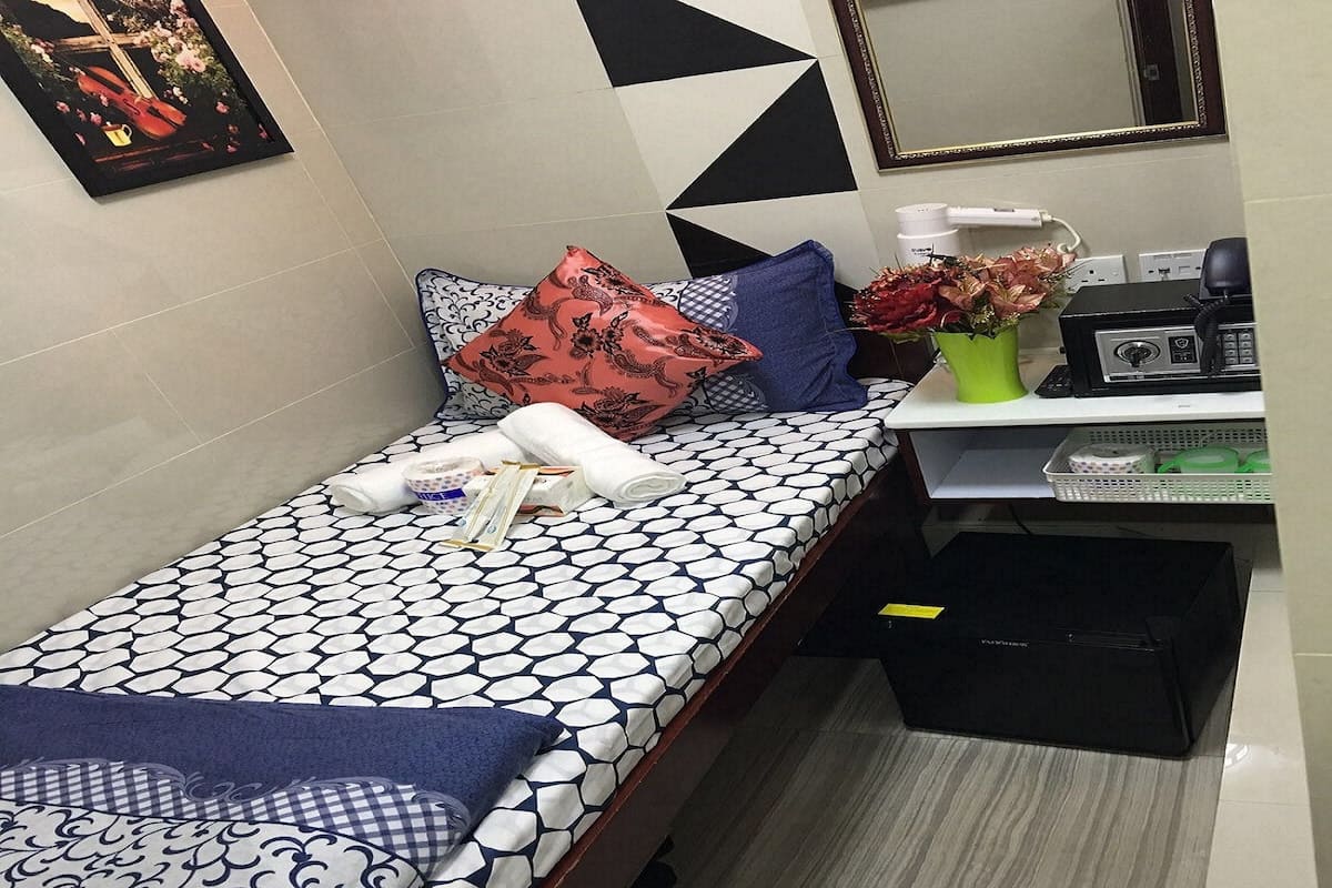 A neatly arranged hotel room with a bed, a small desk, a mirror, and a black trash bin.