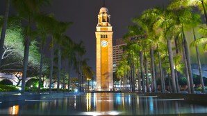 Una alta torre de reloj con esfera, rodeada de palmeras y un estanque reflectante.