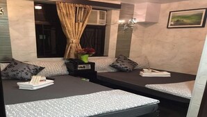 Una habitación con dos camas, cada una con un colchón y almohadas estampadas. Hay una ventana con cortina, una mesita con un florero y un cuadro en la pared.