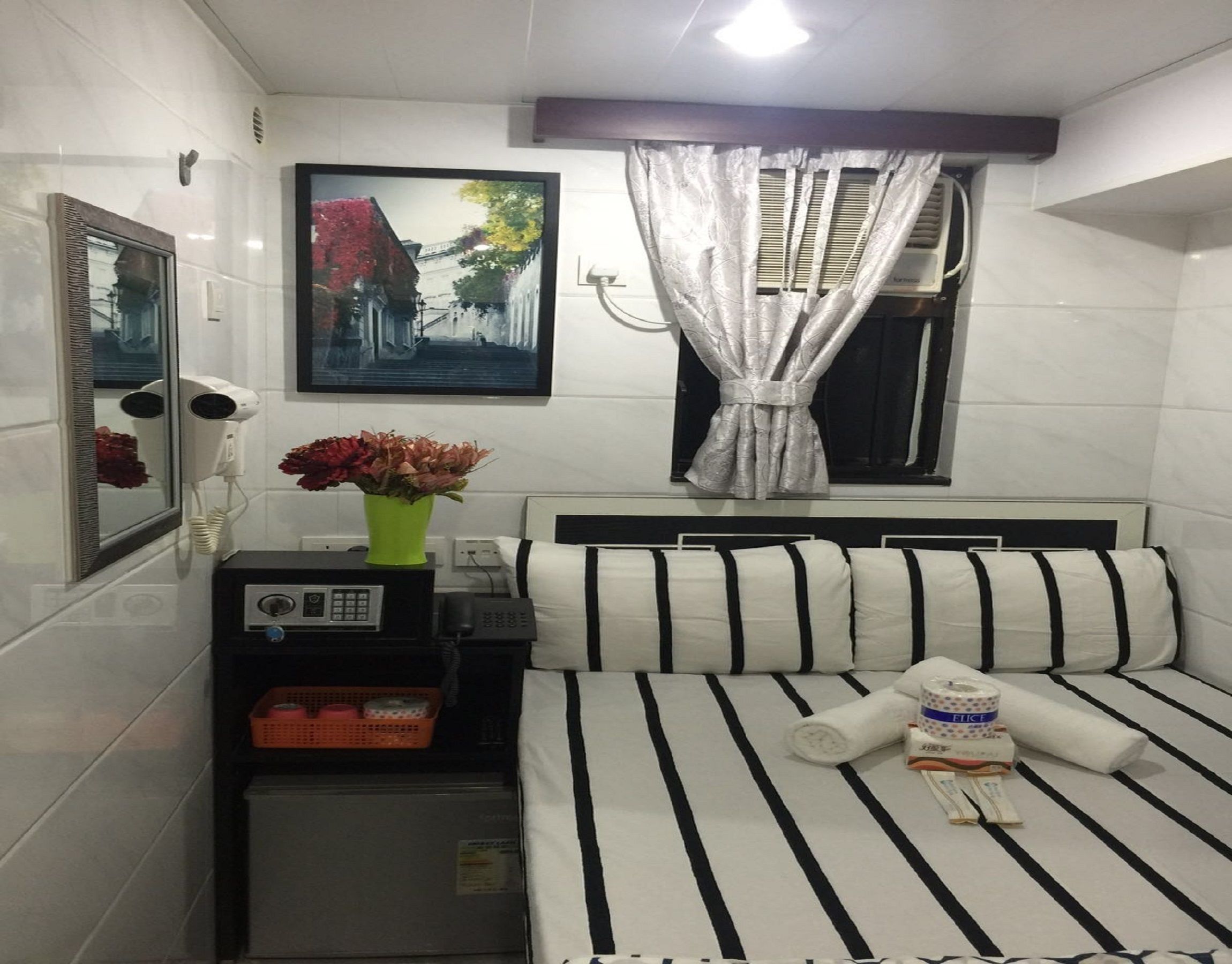 A hotel room with a bed, a small desk with a telephone, a hairdryer, a mirror, a window with curtains, and a wall-mounted picture.