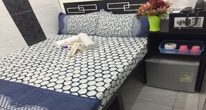A neatly made bed with a patterned bedspread, a small bedside table with a phone and a vase of flowers, and a compact refrigerator. - Skylark Hostel (Kowloon)
