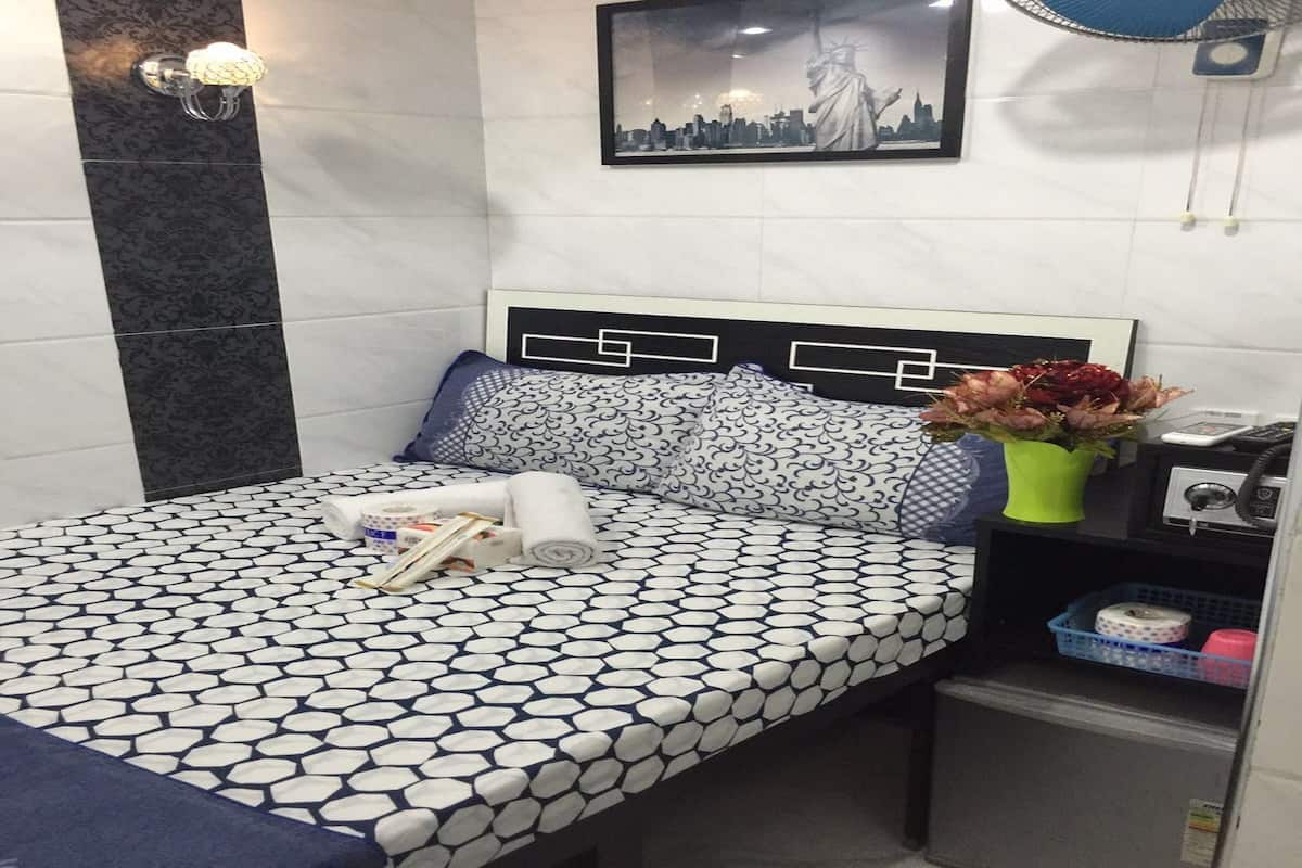 A neatly arranged hotel room with a patterned bedspread, a cityscape artwork, a bedside table with a vase of flowers, and a small storage unit.