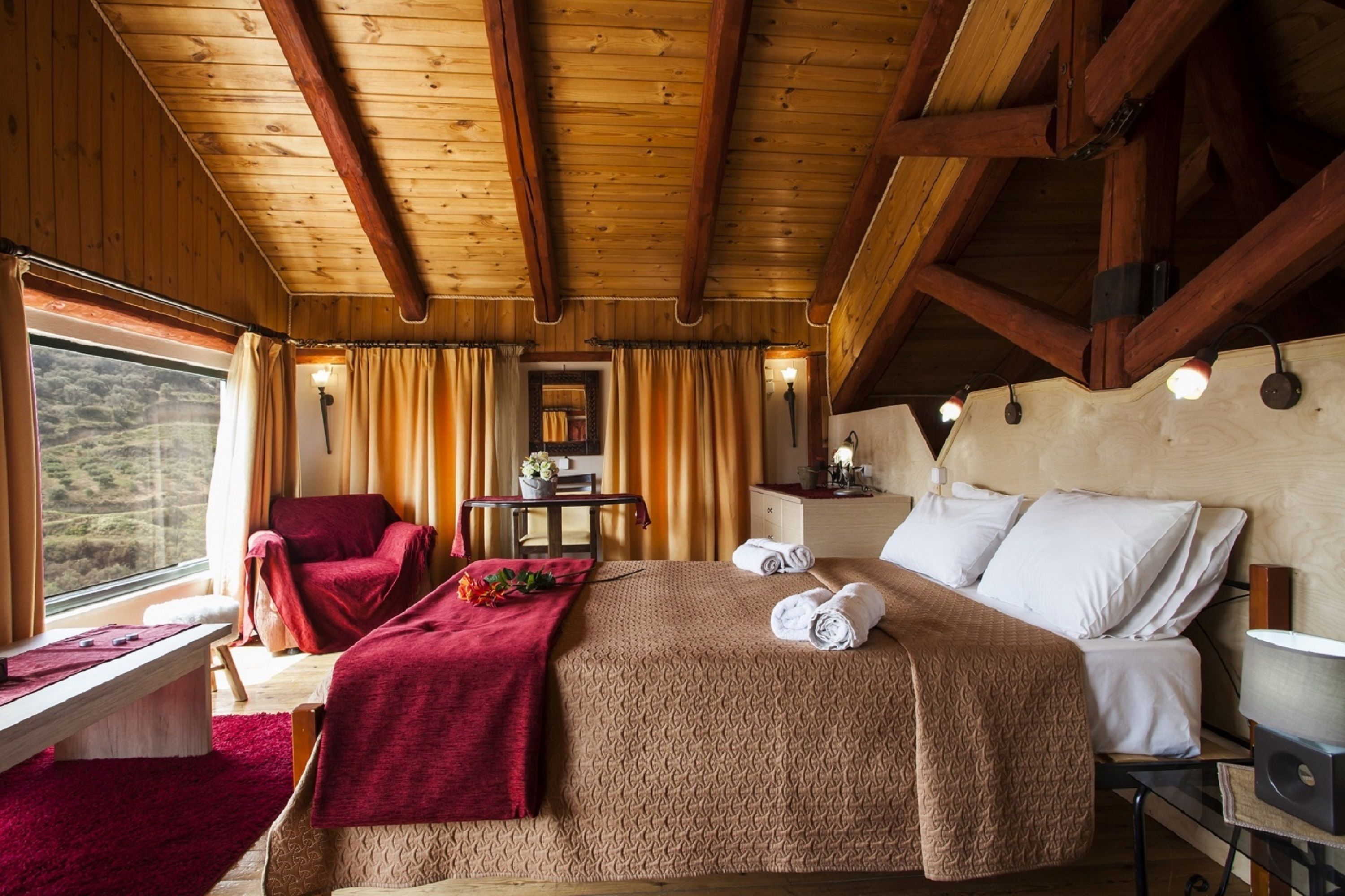 A wooden cabin room with a bed, a red chair, a small table, and a window with curtains.