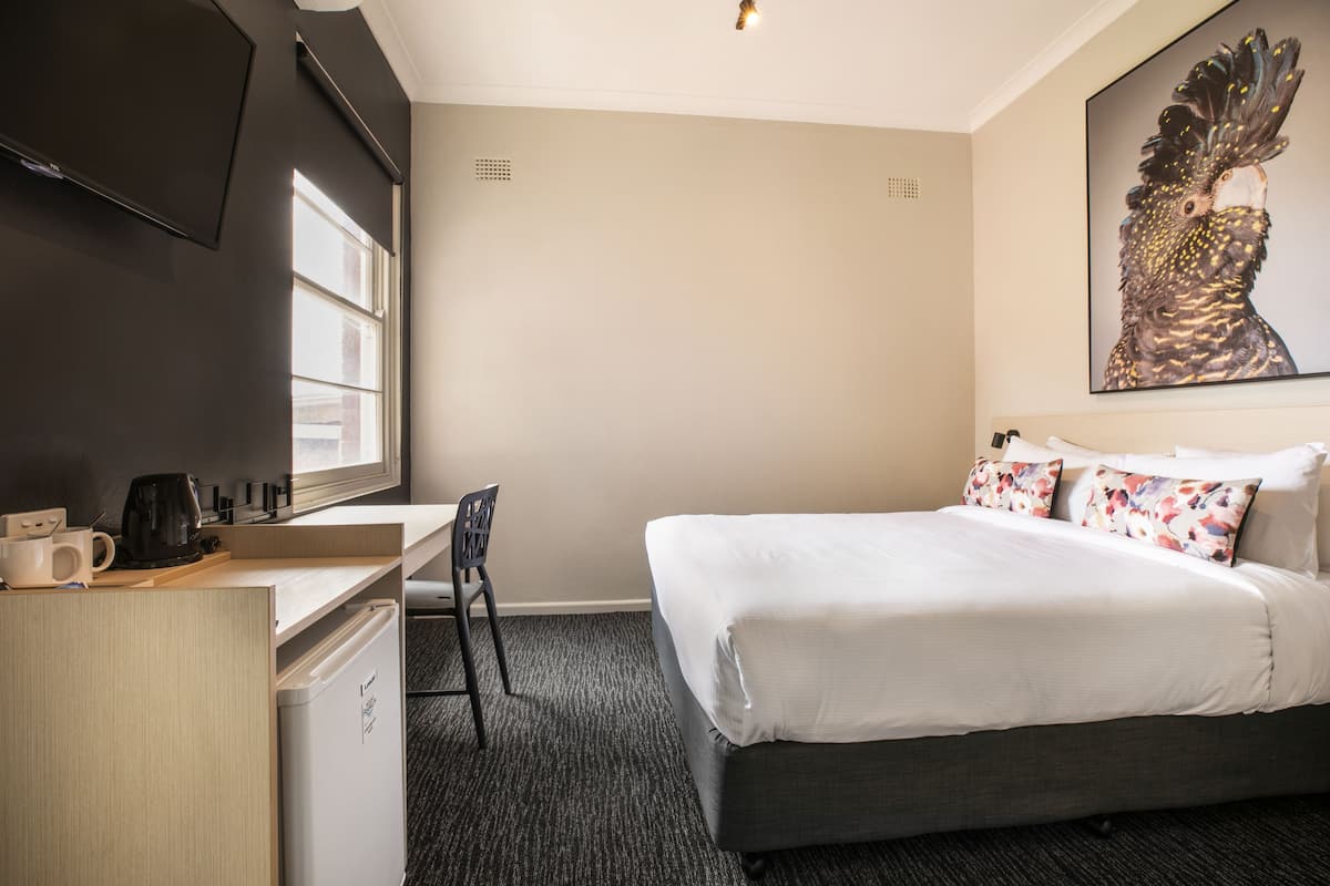 A hotel room with a bed, a desk with a coffee maker, a chair, a TV mounted on the wall, and a painting above the bed.