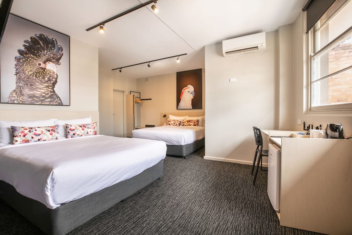 A hotel room with two beds, a desk, and a chair. There are two paintings on the walls, a window, and an air conditioning unit.
