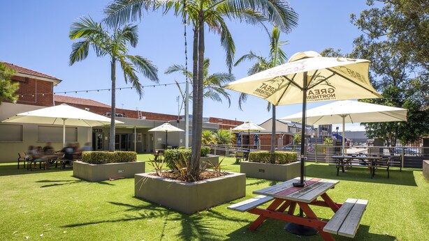 Een terras buiten met picknicktafels en parasols, omringd door palmbomen en plantenbakken.