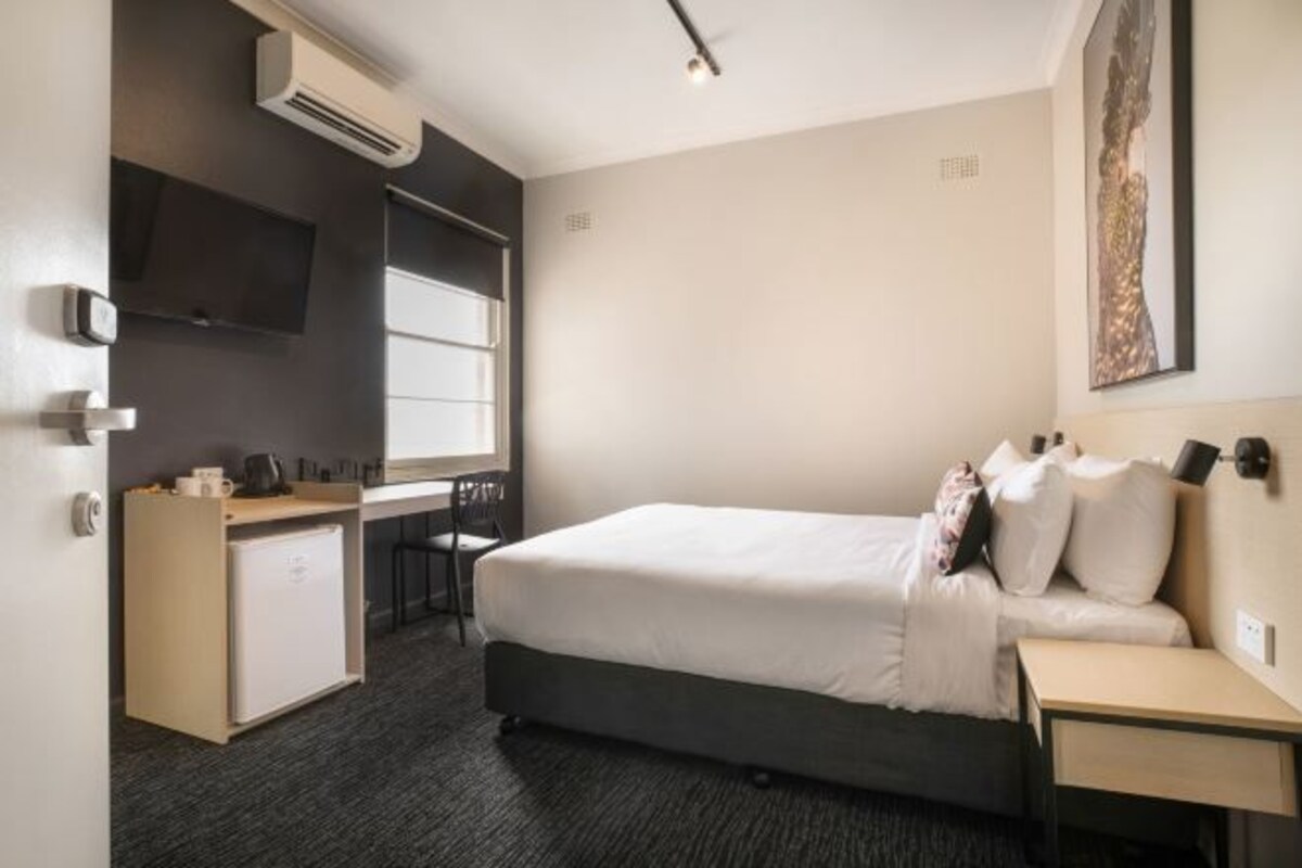 Ett hotellrum med en säng, ett skrivbord, en stol, ett litet kylskåp och en tv som är monterad på väggen.