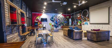 A modern lounge with wooden tables, comfortable seating, a large mural, and a projector screen.