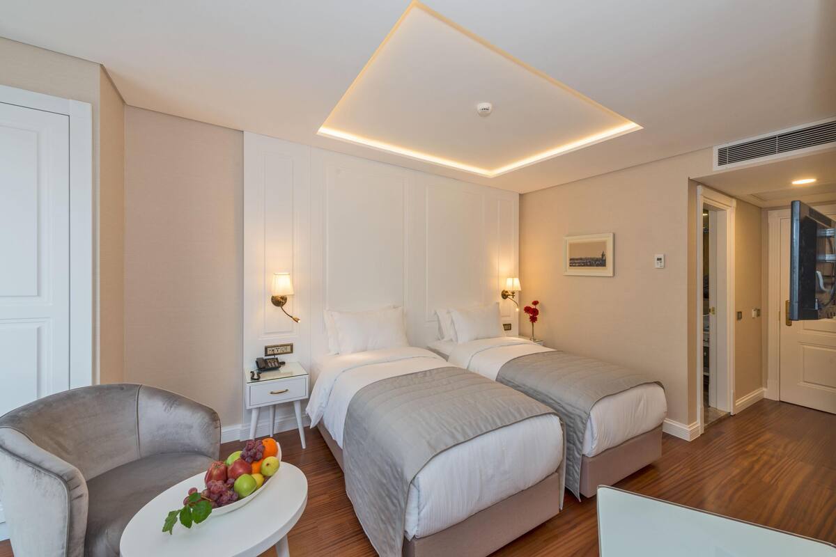 Habitación de hotel con dos camas, una mesita con fruta, una silla y una pared blanca con un cuadro.