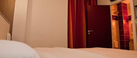 A hotel room with a bed, a door with curtains, a TV mounted on the wall, and a bathroom with red tiles.