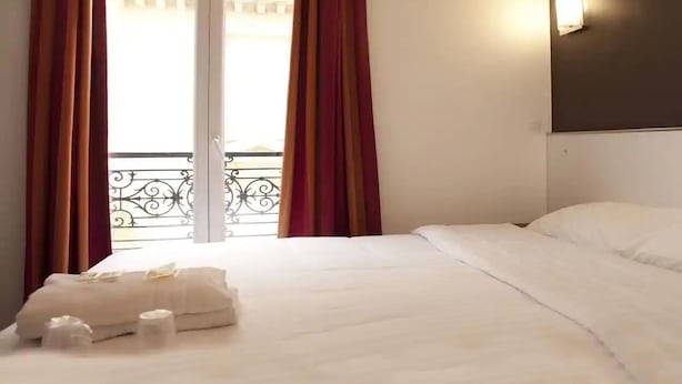 Una camera d'albergo con un letto, una finestra con tende e una lampada a parete, con vista su un edificio.