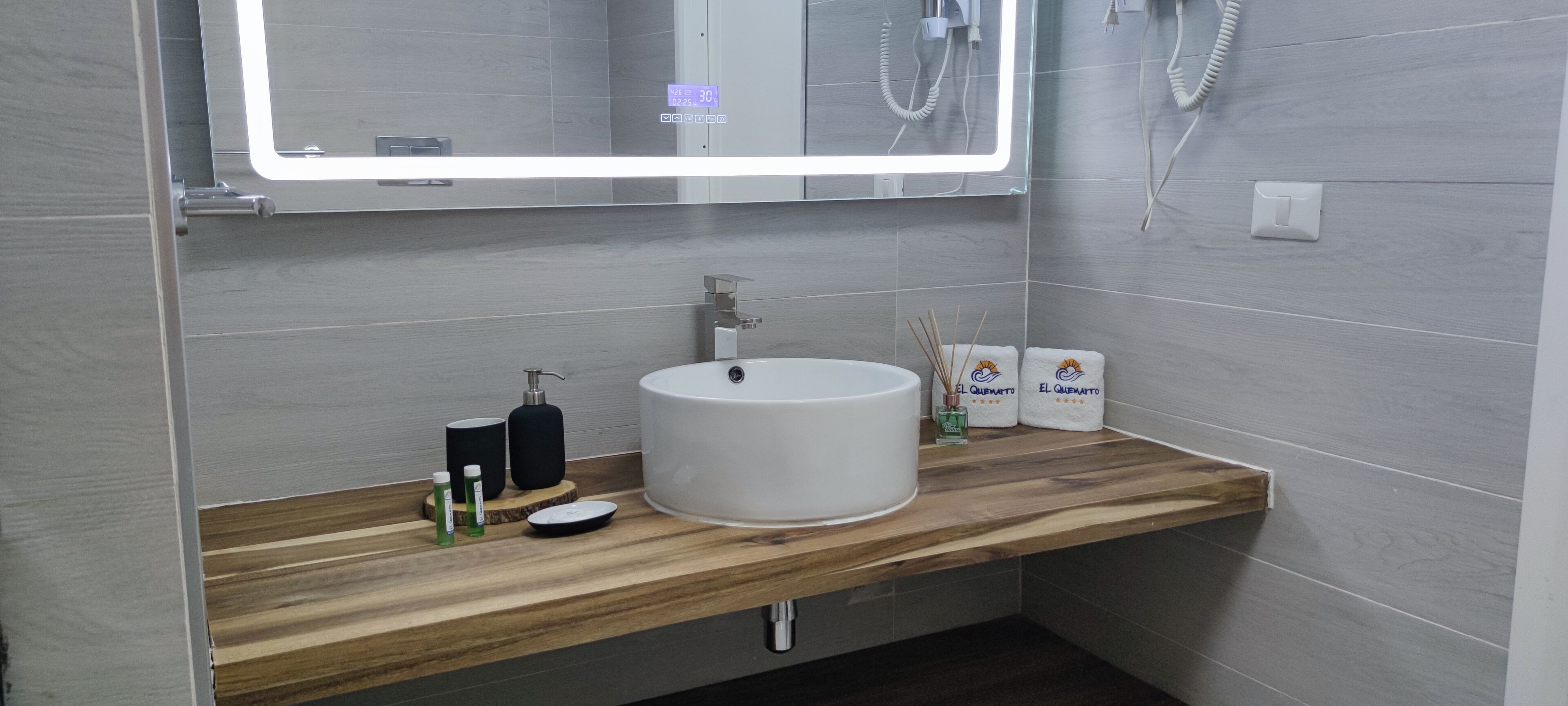 Een moderne badkamer met een houten wastafelmeubel, een witte wasbak en een spiegel met verlichting rondom.