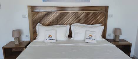 A neatly made bed with two pillows and two towels, each with the logo "El Quenamo".