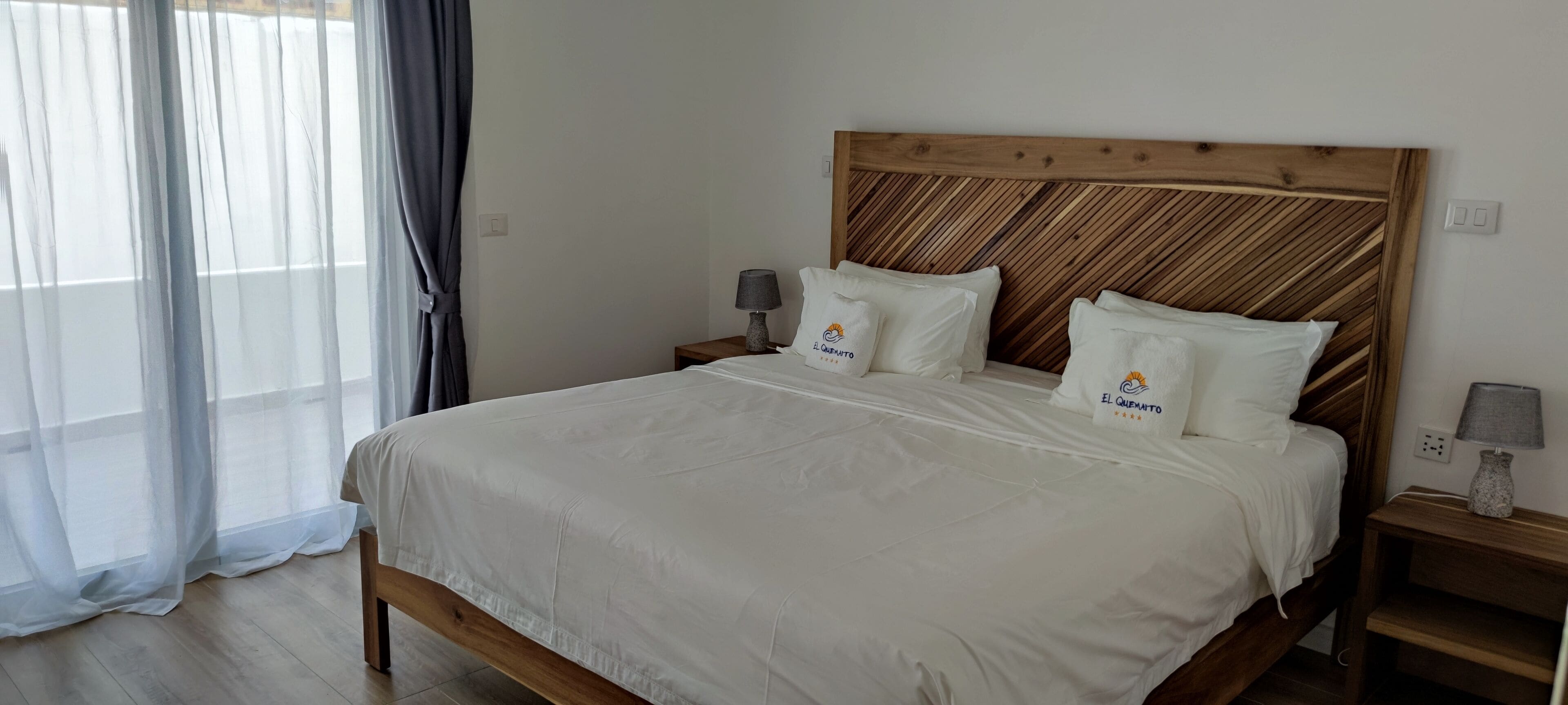 A neatly made bed with a wooden headboard, two bedside tables with lamps, and a window with sheer curtains.