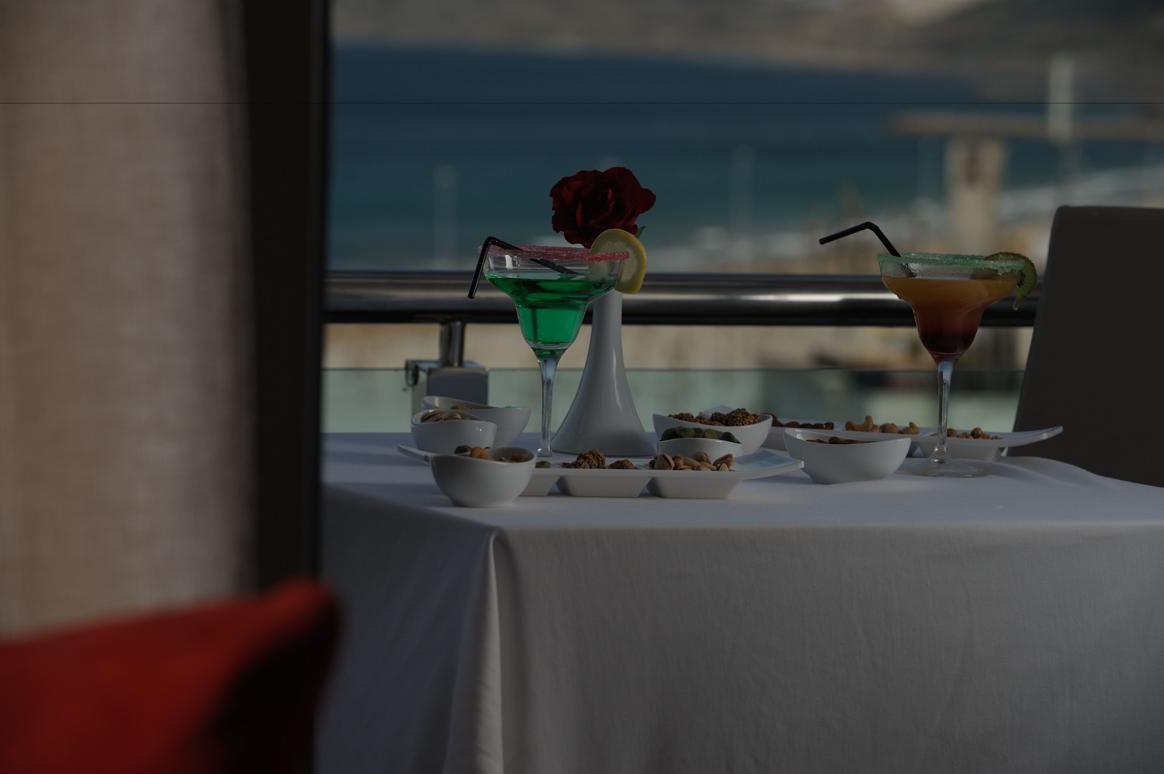 Ein Tisch ist reichhaltig mit Speisen und Getränken gedeckt, eine Vase mit einer Rose steht darauf, und durch ein Fenster ist der Blick aufs Meer gerichtet.