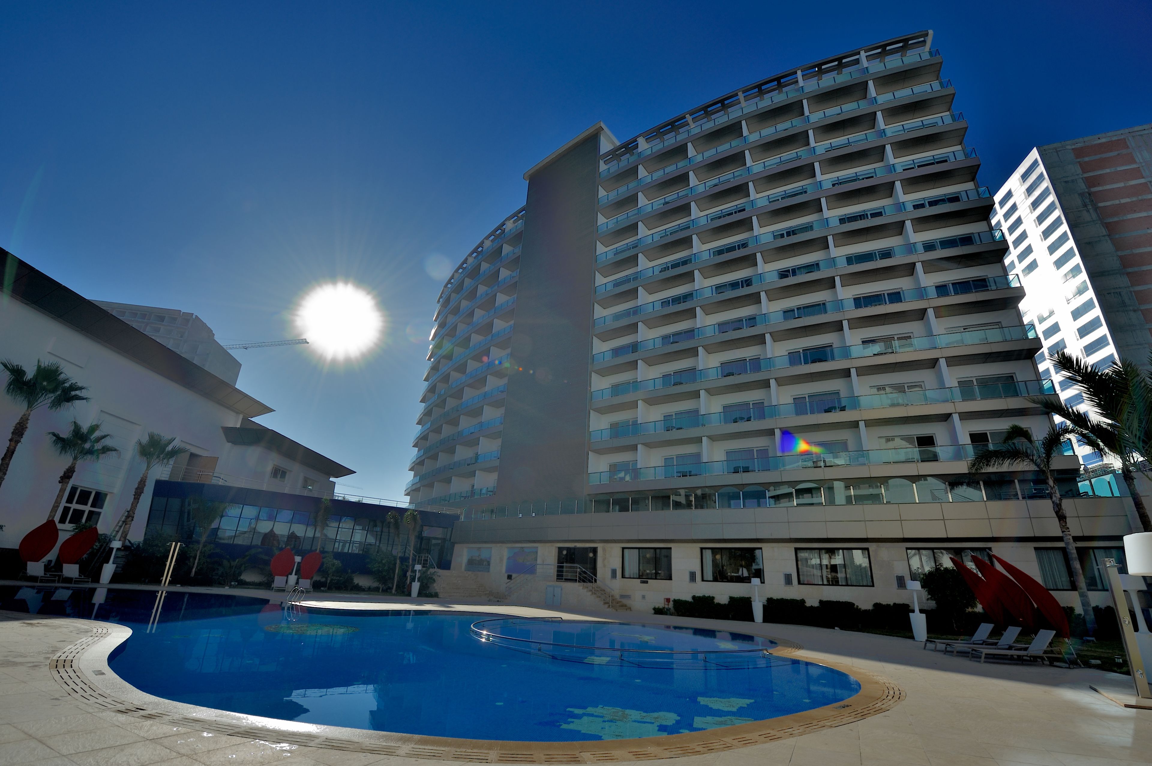 Un hotel moderno con una piscina grande, varios pisos y un cielo azul despejado.