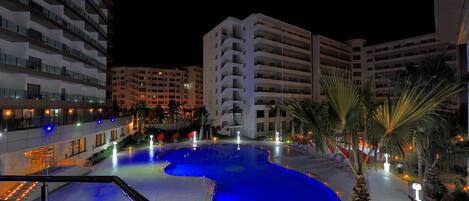 A nighttime view of a hotel with a large swimming pool, multiple floors, and palm trees.