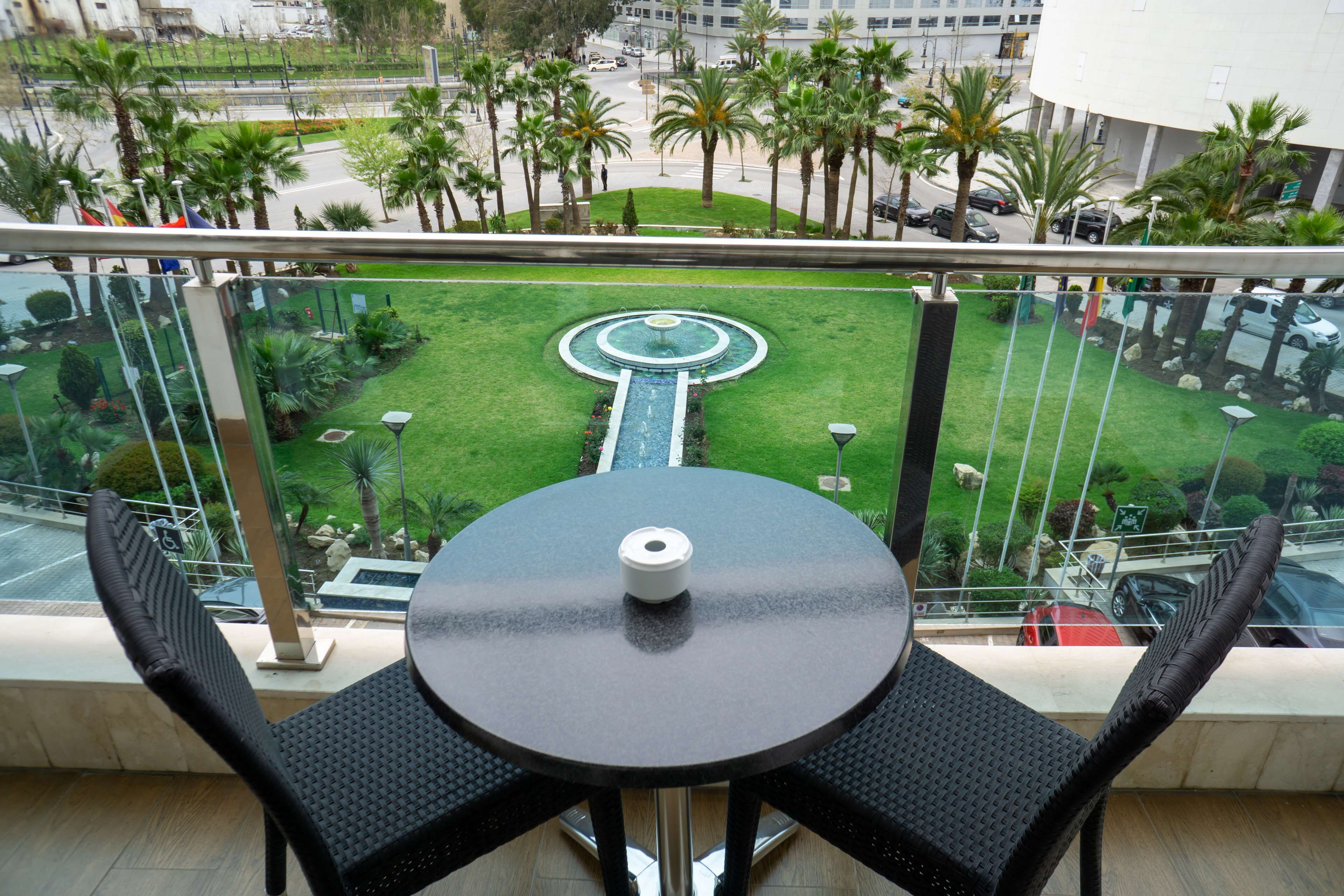 Un balcón con una mesa redonda y dos sillas, con vista a un césped cuidado que incluye una fuente circular y palmeras.
