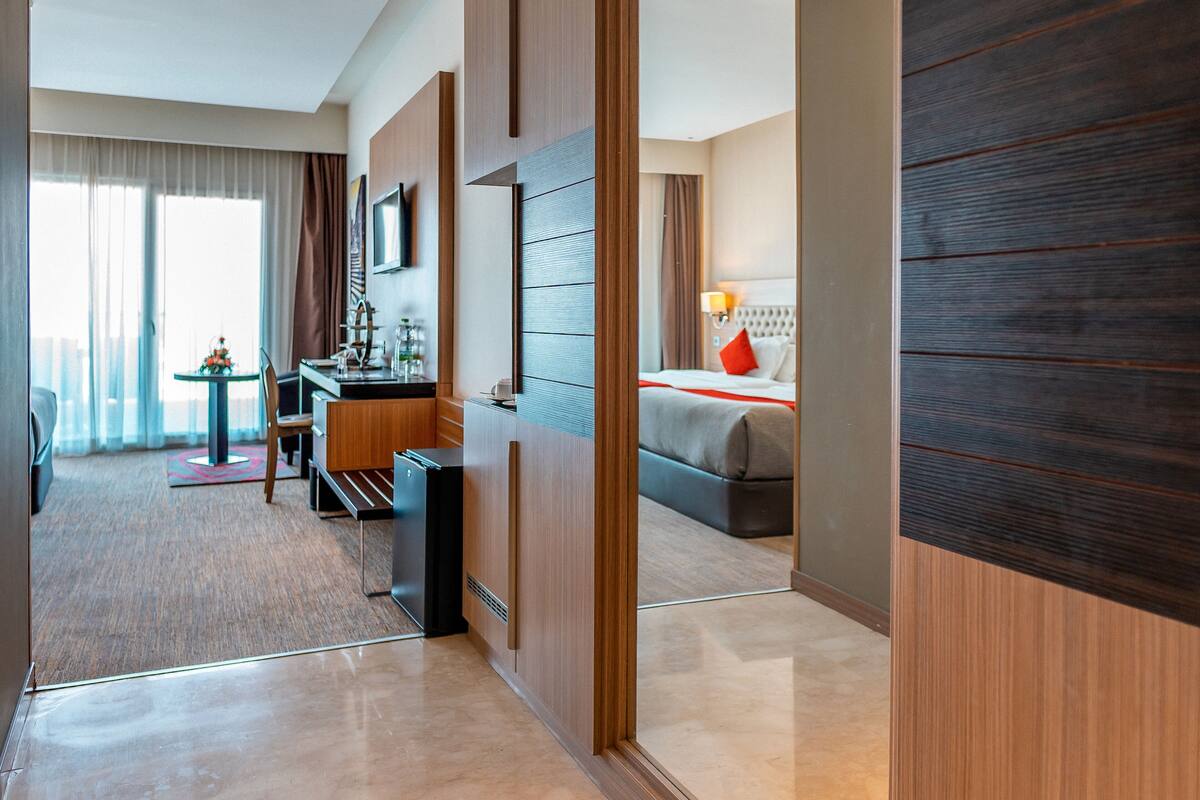 Una habitación de hotel moderna con un armario de madera, una cama con almohadas rojas, un escritorio con una silla y una pequeña mesa con un florero.
