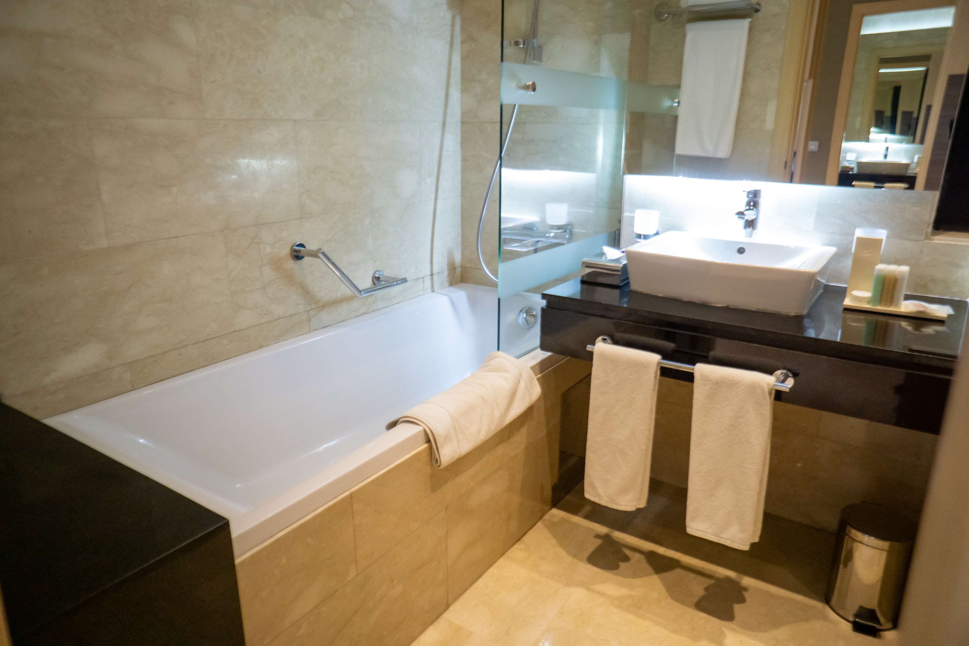 A modern bathroom with a bathtub, a sink, and a mirror. There are towels hanging on the wall and a trash can.