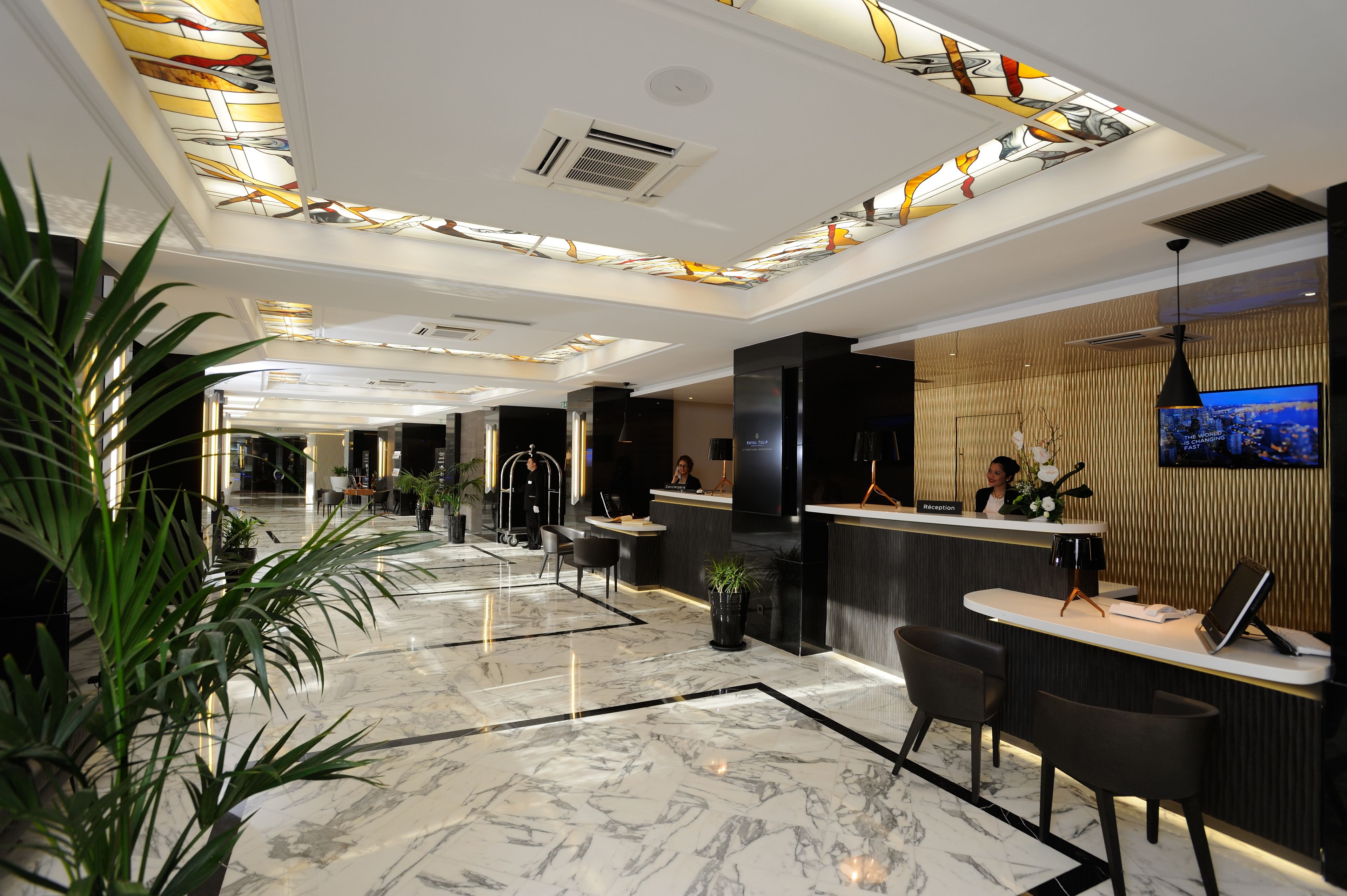 A modern hotel lobby with a reception desk, seating area, and a large TV screen.