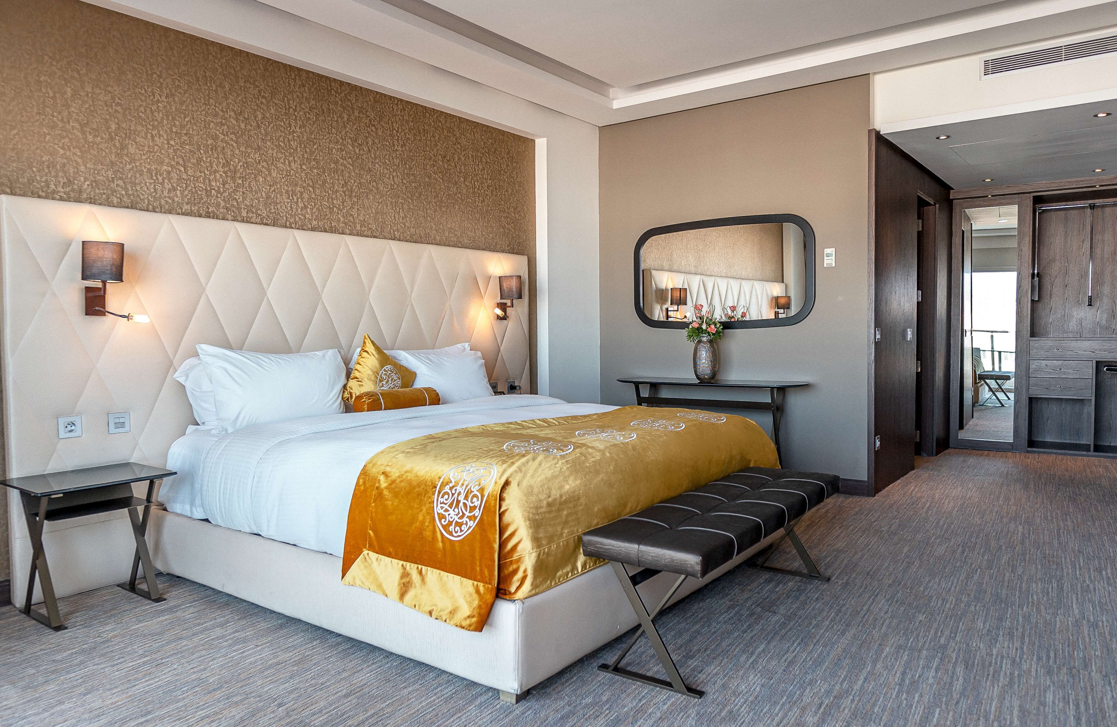 A modern hotel room with a large bed, a bedside table, a bench, a wall-mounted mirror, and a view of the outdoor area.